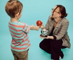 A young boy presents an apple to a crouching woman with a trophy and a teal backdrop.