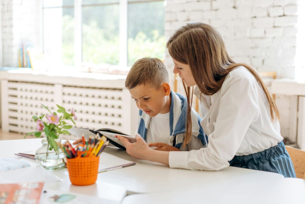 A mother helps her young son read a book in a cozy indoor environment.